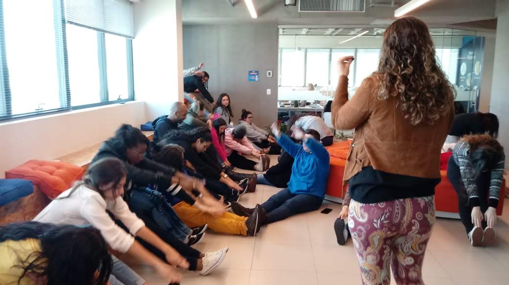 Dani Pinotti conduzindo dinâmica de alongamento durante palestra corporativa, com grupo sentado em roda.