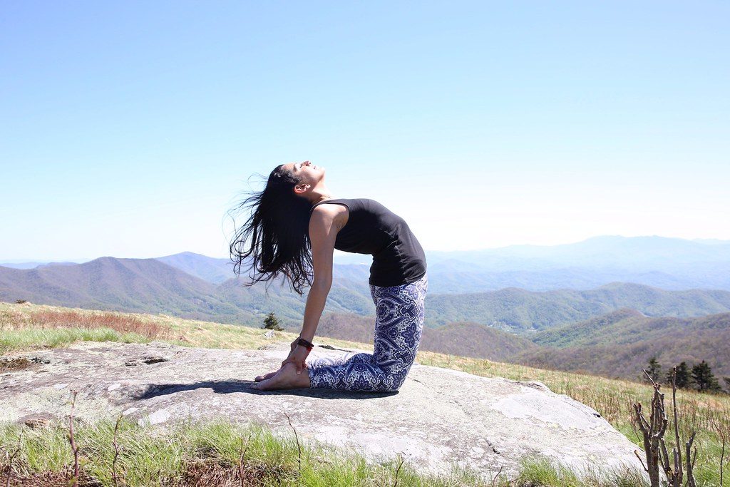 Mulher praticando yoga ao ar livre em meio às montanhas, com os braços erguidos recebendo luz solar, representando bem-estar e respiração consciente.